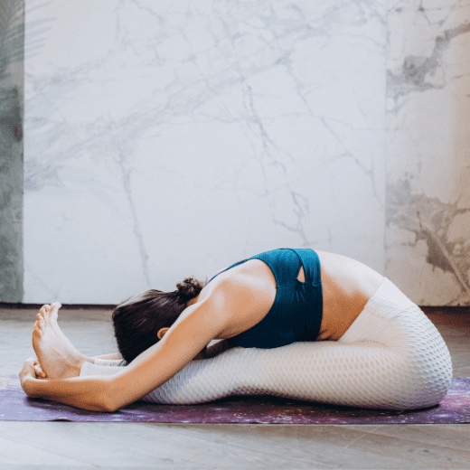 Mulher praticando Paschimottanasana, postura da pinça no yoga, com o tronco alongado sobre as pernas estendidas durante aula presencial no bairro da Tijuca, zona norte do Rio de Janeiro.