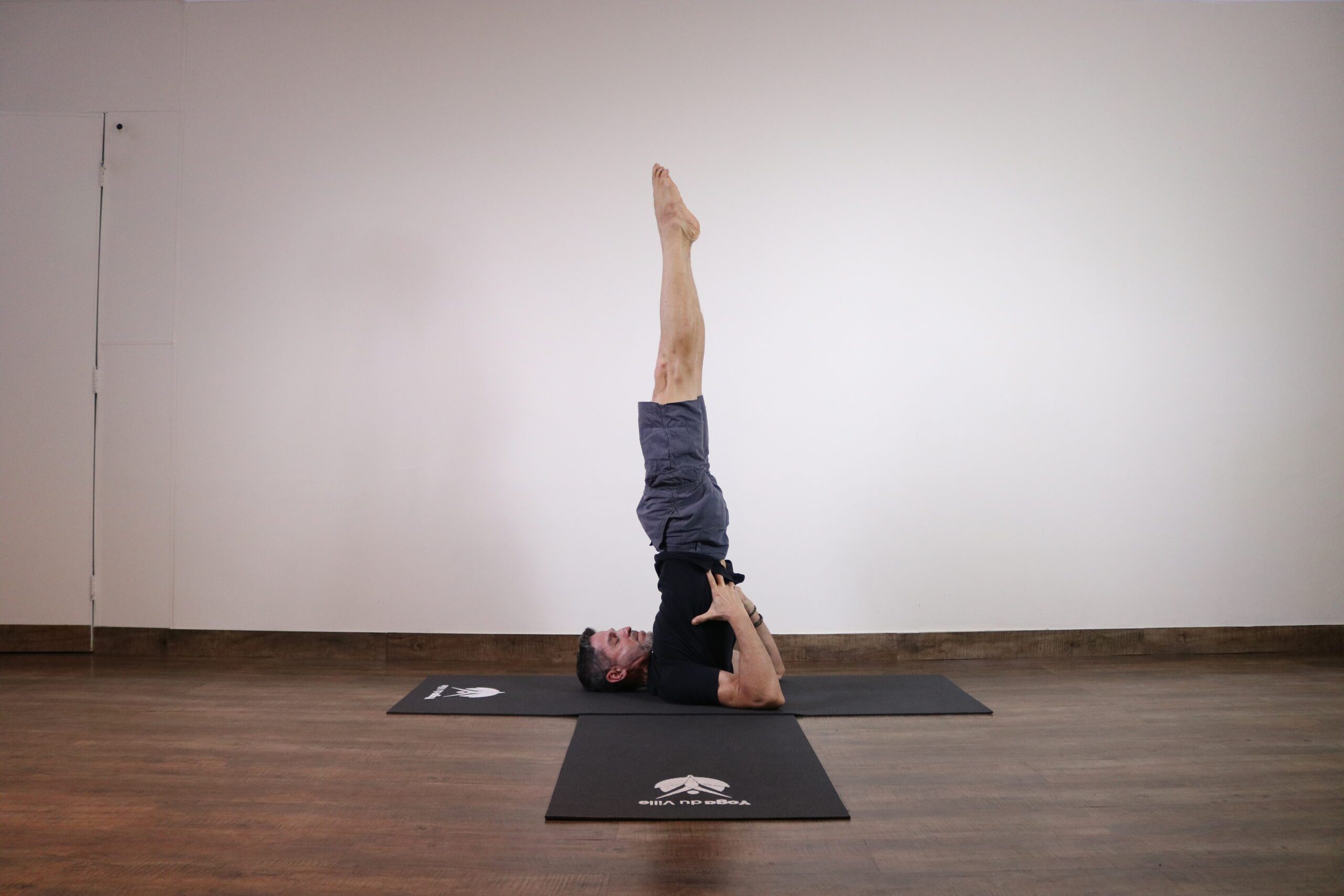 Instrutor Márcio executando Sarvangasana, postura da vela no yoga, durante aula no estúdio Yoga du Ville, Tijuca, RJ